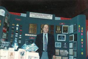 Dave Fertig poses in front of an APS Awards booth at a convention.